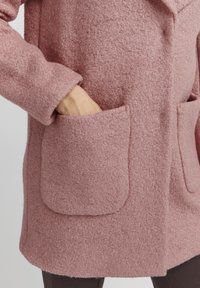 Main partiellement insérée dans une grande poche texturée d'un manteau rose, montrant le tissu épais et doux du manteau ainsi que le design de la poche.