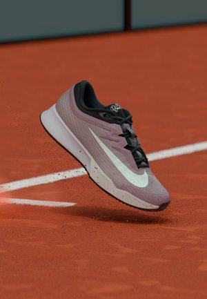 Világoslila és fekete Nike futócipő fehér pipa logóval, amely egy piros agyagos teniszpályáról felemelkedik, fehér határvonalakkal.