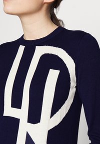 Marinblå stickad tröja med en stor kontrasterande vit grafisk design. Rund krage och texturerade detaljer vid axlarna.