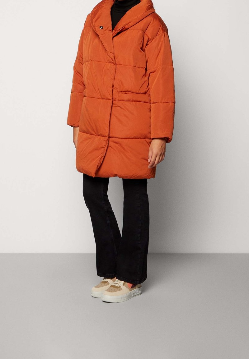 Puffjacka i bränd orange, med en stor krage, quiltat mönster och framknäppning. Matchad med svarta byxor och lätta sneakers.