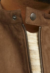 Cuello de una chaqueta de ante marrón con cierre a presión y cremallera de metal. Suéter de punto de color claro visible debajo, con un patrón de rayas.