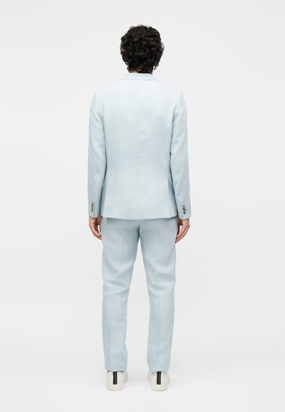 Άτομο με σγουρά σκούρα μαλλιά που φοράει ένα ανοιχτό μπλε κοστούμι και λευκά αθλητικά παπούτσια, στέκεται γυρισμένο με την πλάτη σε ένα απλό λευκό φόντο.