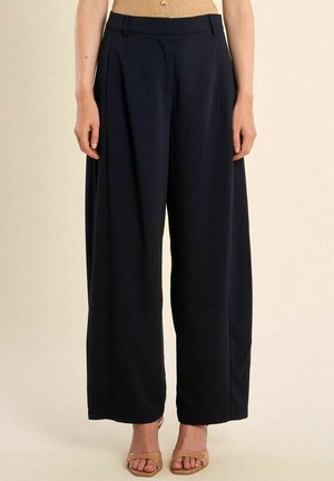 Donna che indossa pantaloni neri a vita alta e gamba larga abbinati a tacchi aperti color nude, in piedi su uno sfondo chiaro e semplice.