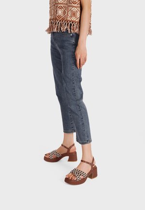 Donna con jeans blu corti, sandali platform intrecciati marroni e bianchi, e una maglia marrone con motivi e frange, in piedi su uno sfondo bianco.