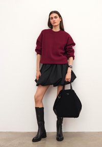 Bordowy dzianinowy sweter, czarna spódnica w kształcie dzwonu, czarne kozaki za kolano i czarna torba na ramię z akcentami. Modelka stoi przy białej ścianie.