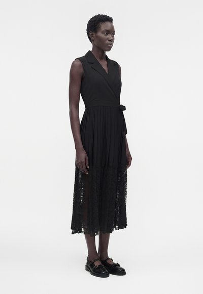 Γυναίκα με κοντά σγουρά μαλλιά, που φοράει αμάνικο μαύρο μίντι φόρεμα με δαντελένιο τελείωμα και μαύρα παπούτσια Mary Jane, στέκεται μπροστά σε λευκό φόντο.