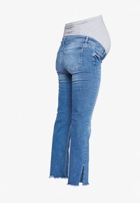 Personne enceinte portant un jean de maternité bleu avec une ceinture élastique et un détail de poche arrière, vue de dos sur fond blanc.