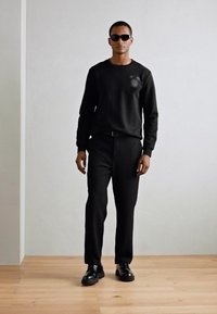 Sweatshirt noir avec un motif texturé et un pantalon noir, complétés par des lunettes de soleil noires et des chaussures noires brillantes, sur un fond neutre.