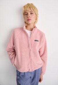 Giovane con capelli biondi corti che indossa una giacca di pile rosa chiaro e pantaloni blu, in piedi su uno sfondo bianco neutro.