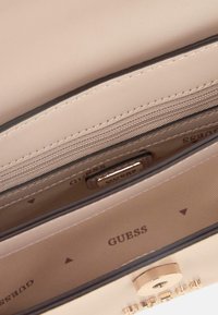 Interno di una borsa in ecopelle beige con una tasca con zip, hardware in tonalità oro e logo "GUESS" stampato sulla fodera in tessuto.
