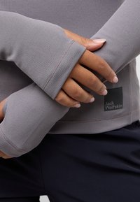 Maglia grigia a maniche lunghe con tessuto testurizzato e a coste, fori per il pollice e un'etichetta nera con logo sul lato, abbinata a pantaloni scuri.