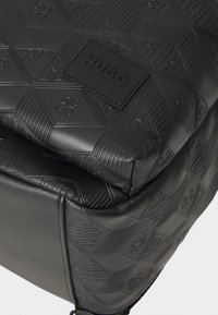 Zaino nero texturizzato con motivo geometrico, marchio in rilievo e finitura in pelle liscia. I dettagli includono una etichetta logo rettangolare.