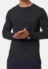 Czarnoszary sweter wykonany na drutach z okrągłym dekoltem, ściągaczami na mankietach i dolnej krawędzi. Gładka tekstura z dopasowanymi rękawami, noszony na granatowych spodniach.