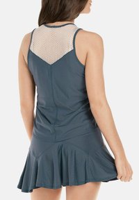 Vrouw in een mouwloze donkerblauwe jurk met een gaaspaneel op de bovenrug en een uitlopende rok, gezien van achteren.