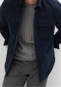 Homme portant une surchemise bleu marine à poches à rabat, un pull gris côtelé et un pantalon gris, debout les bras derrière le dos.