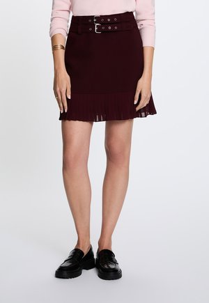 Kvinde iført en mørk burgunderrød nederdel med dobbelt spænde bælte og plisseret kant, lyserød sweater, sorte loafers og gyldne armbånd på håndleddet.