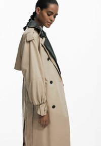 Manteau beige avec col vert surdimensionné, devant double boutonnage, tissu texturé et manches froncées avec accents de boucle.
