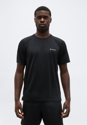 Άνδρας που στέκεται κοιτάζοντας μπροστά, φορώντας μαύρο αθλητικό πουκάμισο Columbia με κοντά μανίκια και μαύρο σορτς, με λευκό φόντο.