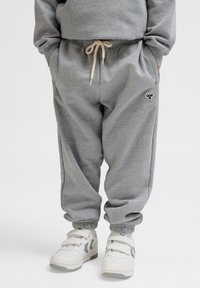 Grijze sweatpants met een elastische tailleband en trekkoord, voorzien van zijzakken en een klein marineblauw logo aan de linkerkant. Gepaard met witte sneakers.