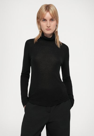 Crna dugih rukava turtleneck majica od mekog, tankog pletiva; sadrži prilagođeni dizajn i minimalistički stil. Kombinirano s tamnim, krojenim hlačama.