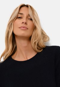 Blondynka z luźnymi falującymi włosami w czarnej bluzie z okrągłym dekoltem, zwrócona lekko w górę z neutralnym wyrazem twarzy na białym tle.