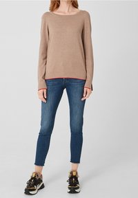 Kobieta ubrana w beżowy sweter z długim rękawem i czerwonym wykończeniem, niebieskie dopasowane dżinsy i czarne oraz beżowe sneakersy z futrzanym wykończeniem, stojąca na jednolitym tle.