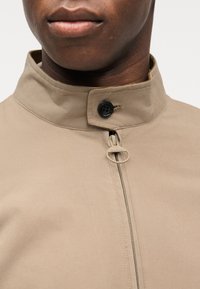 Κοντινό πλάνο ενός ατόμου που φοράει μπεζ σακάκι με ψηλό γιακά, στερεωμένο με μαύρο κουμπί και κεντρικό φερμουάρ με στρογγυ λό τράβηγμα.