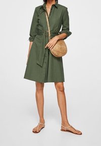 Femme portant une robe chemise ceinturée olive, portant un sac en osier rond en bandoulière, et des sandales plates beiges sur fond blanc.