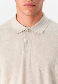 Μπεζ πουκάμισο πόλο με υφή και ανάγλυφο μοτίβο φύλλων. Διαθέτει κλασικό γιακά και πλακέτα με δύο κουμπιά, κατασκευασμένο από μαλακό πλεκτό ύφασμα.