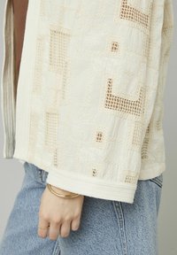 Veste texturée beige clair avec des motifs en dentelle, à devant ouvert et à ourlet nervuré, associée à un denim bleu clair.