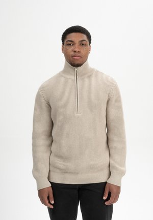 Junger Mann mit kurzen lockigen Haaren und Sommersprossen, der einen beigen Strickpullover mit Viertel-Reißverschluss vorne und schwarze Hosen trägt, stehend vor weißem Hintergrund.