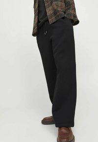 Pantaloni de trening negri, cu o croială lejeră, având o talie elastică și șnururi, asortați cu pantofi din piele maro. Jachetă texturată cu carouri.