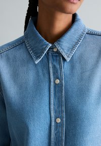 Nahaufnahme einer Person, die ein hellblaues Jeanshemd mit spitzem Kragen und Metall-Druckknöpfen trägt, teilweise Hals und Kiefer sichtbar.