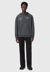 Szary hoodie z przednią kieszenią i detalami logo. W połączeniu z czarnymi spodniami i trampkami. Hoodie ma luźny krój i sznurki do regulacji.