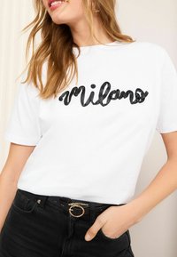 T-shirt en coton blanc présentant un imprimé texturé noir "Milano", associé à un pantalon en denim noir et une ceinture avec des accents dorés.