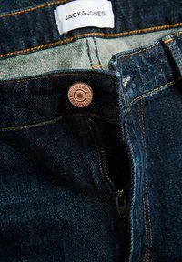 Tumesinised teksapüksid, millel on kontrastsed oranžid õmblused, metallist logo-nupuga ja tõmblukuga esiosa ning silt, millel on kiri "JACK & JONES".