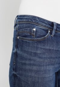 Jeans de mezclilla azul oscuro con corte recto, que presentan bolsillos frontales, costuras sutiles y herrajes metálicos en la cinturilla y los bolsillos.
