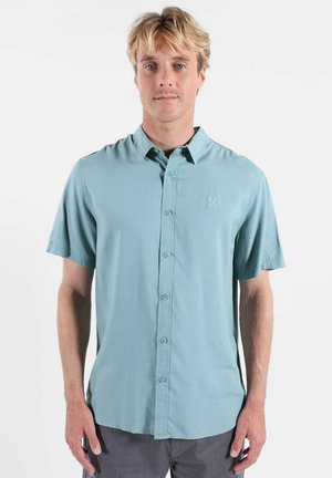 Homme blond portant une chemise à manches courtes bleu clair à boutons et un pantalon gris foncé, debout devant un fond blanc uni.