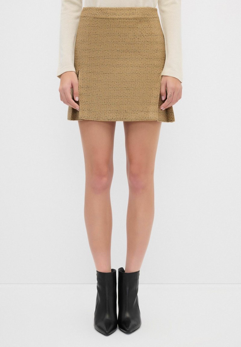 Hame made of textured fabric in a light brown color, featuring a fitted waist and flared hem. Paired with black, pointed-toe ankle boots.