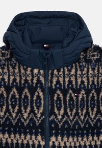 Jachetă din fleece cu capotă matlasată în nuanță de bleumarin, având un model tribal texturat în bej și albastru, cu fermoar frontal. Etichetă cu logo-ul brandului vizibilă.