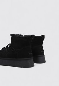 Botines de ante negro con forro de piel sintética, suela de goma plana y lengüeta en la parte trasera. Presentan un diseño minimalista con detalle de cordones.