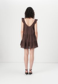 Femme aux cheveux bouclés portant une robe à volants marron foncé sans manches et des talons noirs à brides, debout face à un fond blanc uni.