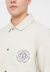 Giacca button-up beige chiaro con colletto a punta, caratterizzata da un logo blu ricamato sulla tasca del petto e bottoni a pressione metallici.