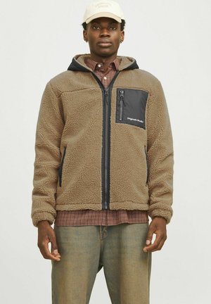 Veste en polaire marron avec une poche avant en nylon noir et une fermeture éclair, dotée d'une capuche et d'une chemise à carreaux en dessous, portée avec un pantalon en jean clair.