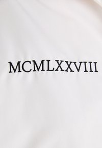 Weißer Stoff mit dem in Schwarz gestickten Text „MCMLXXVIII“. Die Textur wirkt glatt und zeigt ein dezentes Muster.