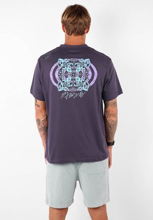 T-shirt gris à manches courtes avec des motifs géométriques colorés dans le dos, porté avec un petit logo sur l'épaule. Associé à un short de couleur claire.