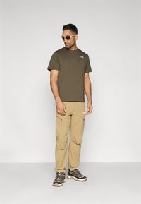 Λαδί κοντομάνικη μπλούζα, χακί cargo παντελόνια με φερμουάρ και σκούρα γκρι παπούτσια πεζοπορίας με ανοιχτές λεπτομέρειες, συνδυασμένα με μπεζ γυαλιά ηλίου.