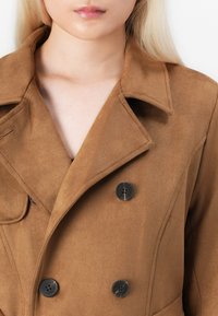 Manteau en suede marron à double boutonnage, avec de grands boutons noirs, des revers crantés et une texture lisse, coupé au niveau de la clavicule.