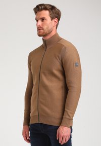 Suéter marrón con cremallera y alto cuello, con detalles acanalados y un parche de logo en la manga. La tela parece suave y texturada.