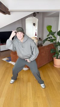 Szürke cipzáras kapucnis pulóver és hozzáillő nadrág, ezüst sportcipők és sötétzöld sapka. Beltéri környezet, fa padlóval és háttérben egy ládás növénnyel.